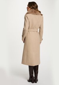 Cappotto in misto lana beige con colletto in pelliccia, maniche lunghe e cintura. Il cappotto presenta un taglio dritto e spacco laterale per facilitare i movimenti.