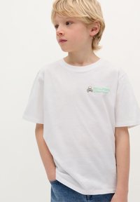 T-shirt bianco in cotone a maniche corte, con un logo a teschio verde e nero sul lato sinistro del petto. Abbinato a jeans in denim di lavaggio medio.