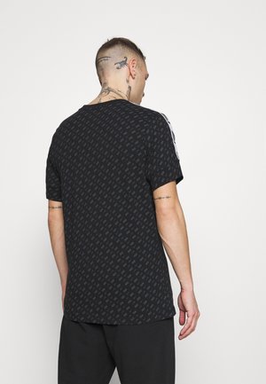 Homme tatoué porte un t-shirt noir à motifs et un pantalon noir, dos à un fond blanc uni.
