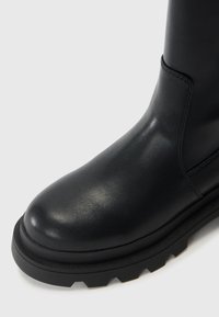 Bota negra de cuero hasta el tobillo con punta redonda y una suela gruesa y texturizada. Presenta costuras elegantes y un acabado liso y brillante.