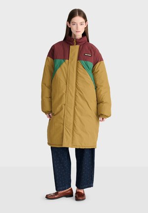 Jeune femme debout portant un long manteau matelassé color block marron, bordeaux et vert avec un pantalon foncé et des chaussures marron.