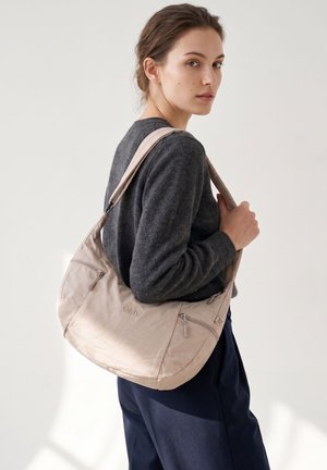 Vrouw die een donkergrijze trui en marineblauwe broek draagt, met een lichtbeige schoudertas met ritszakken.