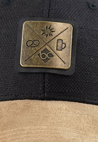 Black cap with a tan suede brim featuring a rectangular bronze logo patch. The patch displays four symbols: a sun, pretzel, beer mug, and "bc" text.