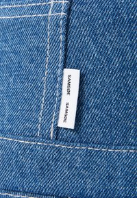 Denim tkanina u plavoj nijansi s vidljivim uzorkom tkanja. Akcentirana bijelim šavovima i etiketom s natpisom "SAMSOE SAMSOE."