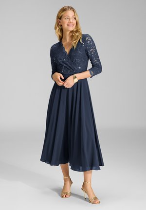 Femme portant une robe en dentelle bleu marine avec des manches trois-quarts et une jupe fluide, accessoirisée d'un bracelet en or et des talons, souriant de côté.