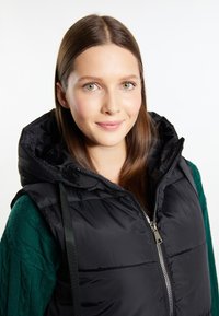 Junge Frau mit hellgrünen Augen und braunen Haaren trägt eine schwarze Daunenweste mit Kapuze über einem dunkelgrünen Strickpullover und lächelt dezent.