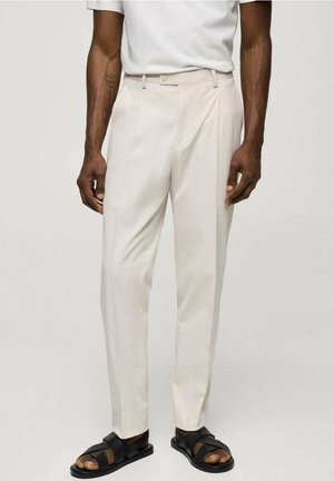 Man draagt lichtbeige op maat gemaakte broek, wit overhemd en zwarte open-toe sandalen, staand tegen een effen achtergrond.