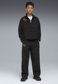 Svart träningsoverall med röda accenter, bestående av en jacka med dragkedja och lösa byxor. Inkluderar Puma-logotyp och en rund logotyp på jackan.