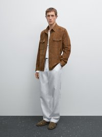 Braune Wildlederhemdjacke mit zwei Brusttaschen, darunter ein weißes Hemd, lockere weiße Hose und olivgrüne Sandalen mit Ausschnitten.