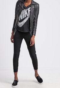 Person klädd i svart läderjacka, svart Nike t-shirt med grafiskt tryck, svarta byxor och svarta slip-on skor, står mot en enkel bakgrund.