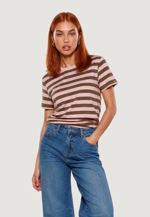 Donna con i capelli rossi che indossa una maglietta a righe rosa e marrone e jeans blu, in piedi con una mano in tasca.