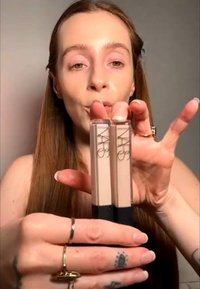 Donna con capelli lunghi che tiene due tubetti di correttore liquido NARS, mostrandoli vicino alla fotocamera per confronto.