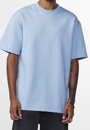 Mężczyzna w jasnoniebieskiej koszuli z krótkim rękawem z haftowanym napisem "NN.07" na klatce piersiowej, czarnych dżinsach oraz z widocznymi tatuażami na prawym przedramieniu.