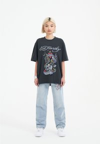 T-shirt nera oversize con grafica colorata raffigurante un teschio e ali, abbinata a jeans blu chiaro con ricami floreali.