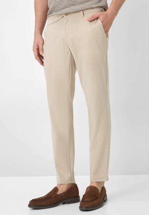 Man wearing beige slim-fit trousers and brown suede loafers, hand in pocket, against a plain white background.