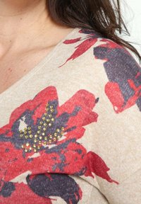 Beżowy sweter z czerwonym i granatowym kwiatowym wzorem, z dodatkowymi akcentami w postaci złotych cyrkonii na wzorze kwiatowym. Miękka, teksturowana tkanina.
