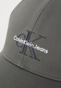 Oliven grønn baseballcaps laget av stoff, med en strukturert design. Brodert med "CalvinKleinJeans" og "CK" i kontrasterende trådtyper.