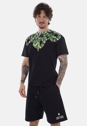 Hombre con cabello rizado y tatuajes lleva un conjunto negro con gráfico de serpiente verde en el escote de la camisa, de pie con una mano en el bolsillo.