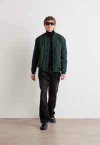 Jachetă bomber verde peste un pulover negru cu guler înalt, combinată cu blugi negri și ochelari de soare, mergând pe un pardoselă gri deschis.
