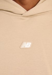 Felpa beige con una texture morbida, caratterizzata da un'applicazione in gomma bianca con il logo sulla parte sinistra del petto. Il tessuto presenta un leggero motivo mélange.