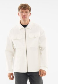 White jacket with a zip closure, two chest pockets, and a classic collar. The fabric has a smooth texture; worn over a black shirt.