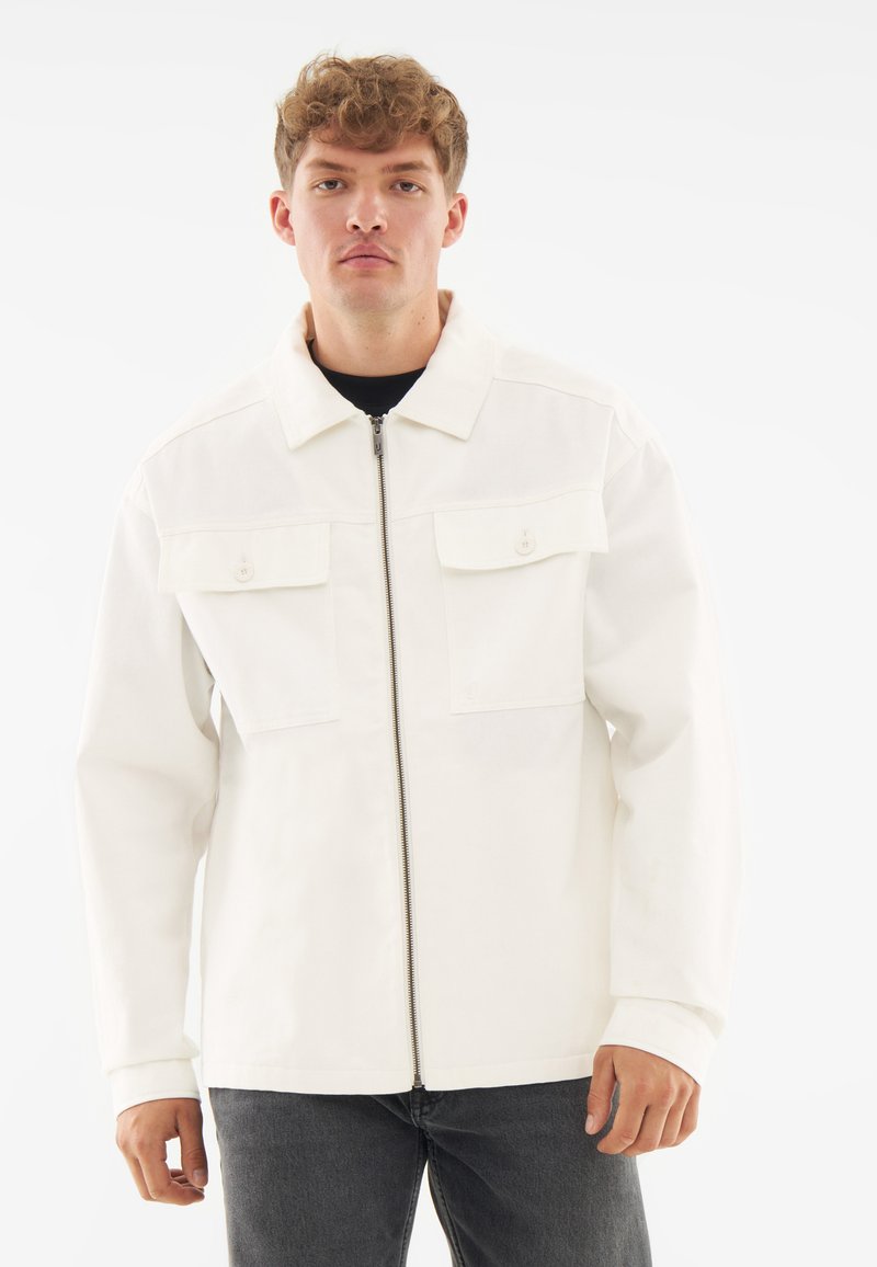 White jacket with a zip closure, two chest pockets, and a classic collar. The fabric has a smooth texture; worn over a black shirt.