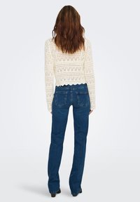 Pull court en maille blanche avec motifs ajourés et ourlet festonné, associé à un jean évasé bleu foncé avec poches arrière et coupe classique.