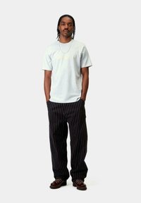 Camiseta gris con un logo amarillo, combinada con pantalones negros a rayas. El modelo se presenta con confianza, usando zapatos marrones. Look general casual.