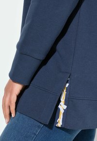 Námořnická modrá mikina s ribbed manžetami, s bočním rozparkem, který odhaluje vzorované vnitřní podšívání v žluté a bílé barvě. Měkká textilie.