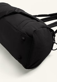 Geantă padded neagră cu formă rectangulară, suprafață texturată, curele ajustabile și picioare circulare la bază pentru stabilitate.