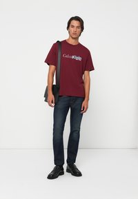 Maroon cotton t-shirt with "Calvin Klein" logo in white and blue, paired with dark jeans and black shoes, holding a black shoulder bag.