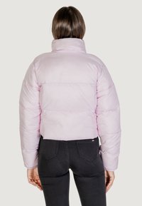 Giacca puffer cropped di colore rosa chiaro con colletto alto e sezioni cucite. Indossata sopra pantaloni neri aderenti, che mostrano una texture liscia.