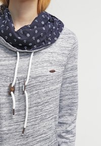 Žena v šedé melírované mikině s bílými šnůrkami a námořnickým šálou s bílými kotvami a kormidly.