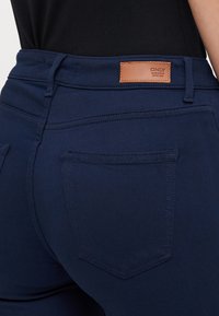 Mørkeblå denimjeans med en tettsittende design, baklommer og en brun lærmerke på linningen. Glatt tekstur.