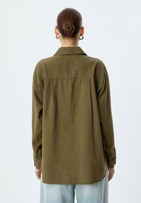 Camicia verde oliva a maniche lunghe con bottoni, dal taglio rilassato, texture semplice e dettaglio della piega sul retro. Vista di schiena.