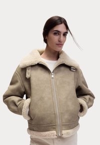 Beige schapenvacht gevoerde jas met een ritssluiting aan de voorkant, grote kraag en twee zijzakken. Zachte textuur en een comfortabele pasvorm, met metalen hardware accenten.
