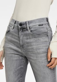 Jeans de denim gris con corte ajustado, que cuentan con bolsillos frontales, un sutil desvanecimiento y una etiqueta negra de G-Star en la cinturilla. Parte superior blanca acanalada visible.
