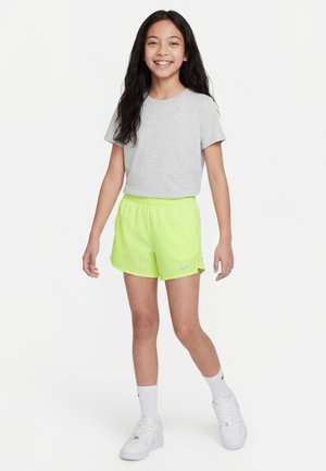 Uśmiechnięta dziewczyna z długimi ciemnymi włosami, ubrana w jasnoszary t-shirt, neonowożółte spodenki Nike, białe skarpetki i białe trampki, stojąca na białym tle.