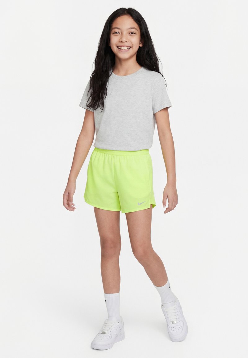 Fille souriante aux longs cheveux foncés portant un t-shirt gris clair, un short Nike jaune néon, des chaussettes blanches et des baskets blanches, debout sur un fond blanc.