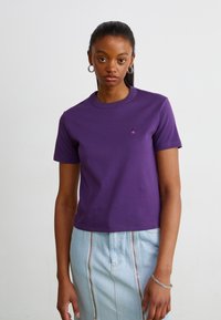 Camiseta de algodón morada con cuello redondo y mangas cortas, que presenta un pequeño logo en el pecho. Combinada con una falda azul clara.