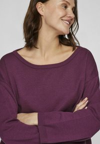 Pull en maille violette avec décolleté large et épaules tombantes. Texture douce et poignets côtelés. Le mannequin se tient avec les bras croisés.