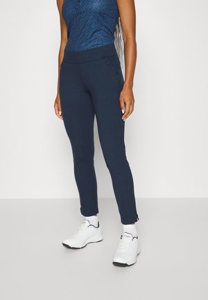 Mujer con pantalones ajustados azul marino, blusa sin mangas con estampado, calcetines blancos y zapatillas deportivas blancas, de pie contra un fondo claro y liso.