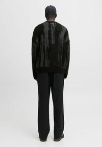 Mann mit einem schwarzen und grauen gemusterten, flauschigen Pullover, schwarzen weiten Hosen, schwarzem Mütze und dunklen Schuhen, der vor einem schlichten Hintergrund von hinten steht.