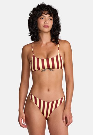 Woman with curly dark hair wearing red and yellow vertical striped bikini, standing against plain light background.