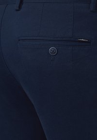 Granatowe spodnie z teksturowanej tkaniny, tylną kieszenią z zapięciem na guzik oraz małą metką marki.