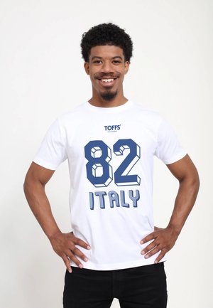 Lächelnder Mann mit lockigem Haar, der ein weißes T-Shirt mit "TOFFS", der Nummer "82" und "ITALY" in Blau trägt und mit den Händen in der Hüfte steht.