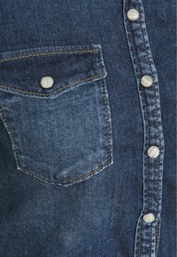 Gros plan sur une chemise en denim montrant une poche poitrine avec rabat et boutons-pression en métal le long de la patte de boutonnage avant.