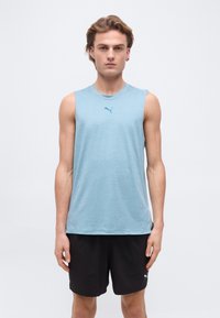 Νέος άνδρας που φοράει ανοιχτό γαλάζιο αμάνικο μπλουζάκι Puma και μαύρο σορτς Puma, στέκεται μπροστά σε απλό φόντο.