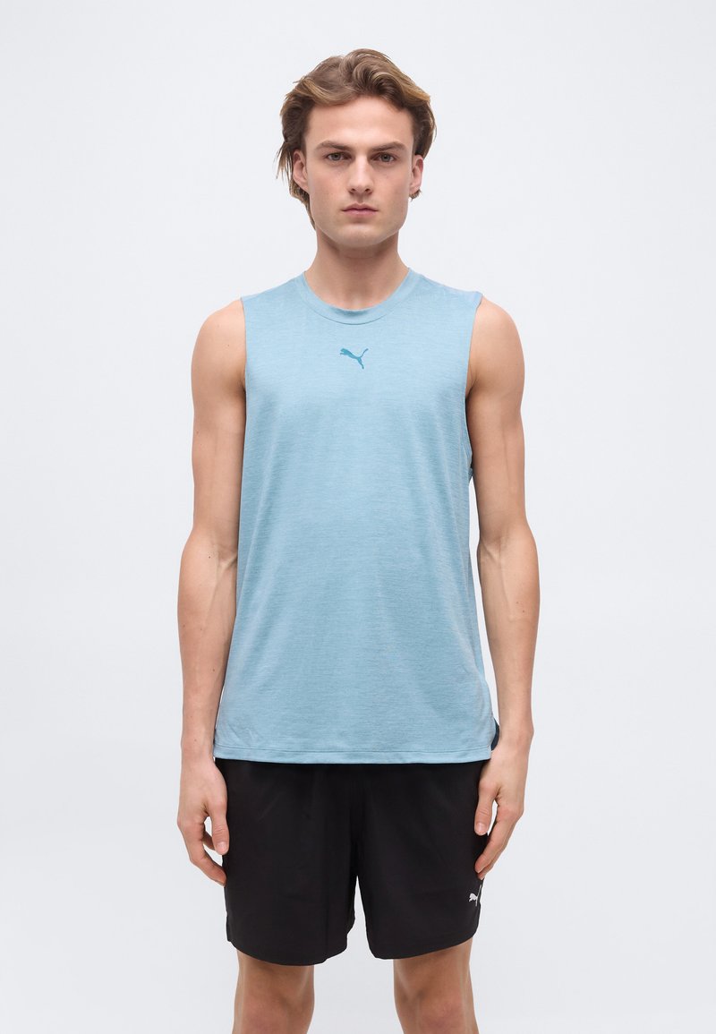 Νέος άνδρας που φοράει ανοιχτό γαλάζιο αμάνικο μπλουζάκι Puma και μαύρο σορτς Puma, στέκεται μπροστά σε απλό φόντο.