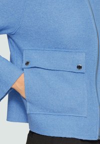 Niebieska kurtka z wełny z zamkiem z przodu, z oversize'owymi rękawami i kieszenią na zatrzask. Teksturowany materiał o miękkim wykończeniu.